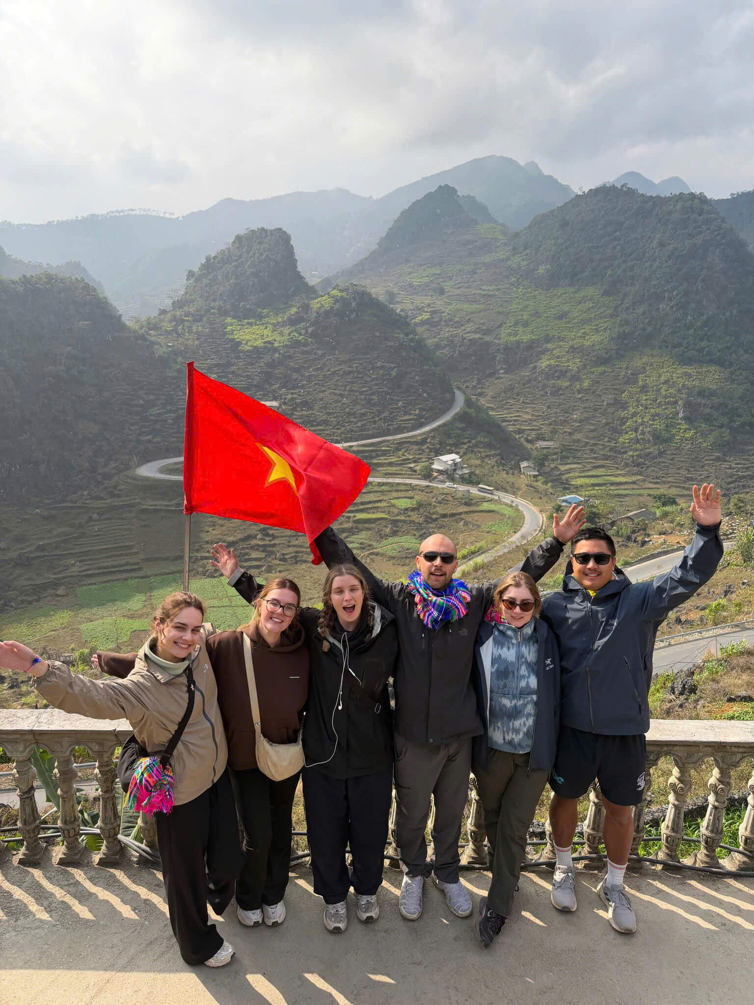 A group of happy travelers holding the Vietnam flag at a scenic viewpoint, showcasing the best time to visit Ha Giang Loop with clear skies and mountain views.